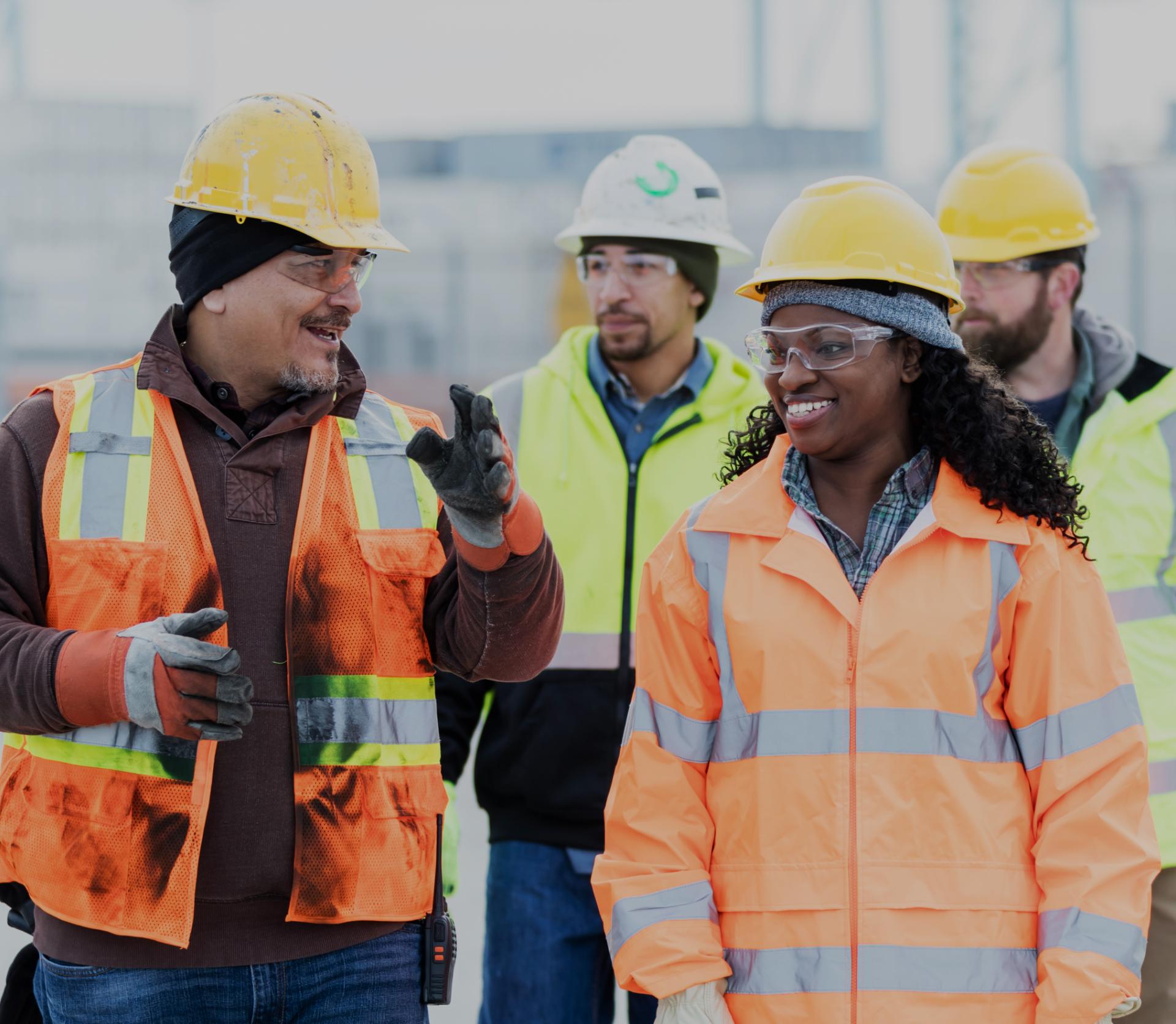A group of construction workers, equipped with hard hats and high-visibility vests, are collaborating at a large industrial job site surrounded by steel structures and heavy equipment. This scene represents the incredible workforce engaged in skilled trades, contributing to building America's future and enhancing the United States labor market.